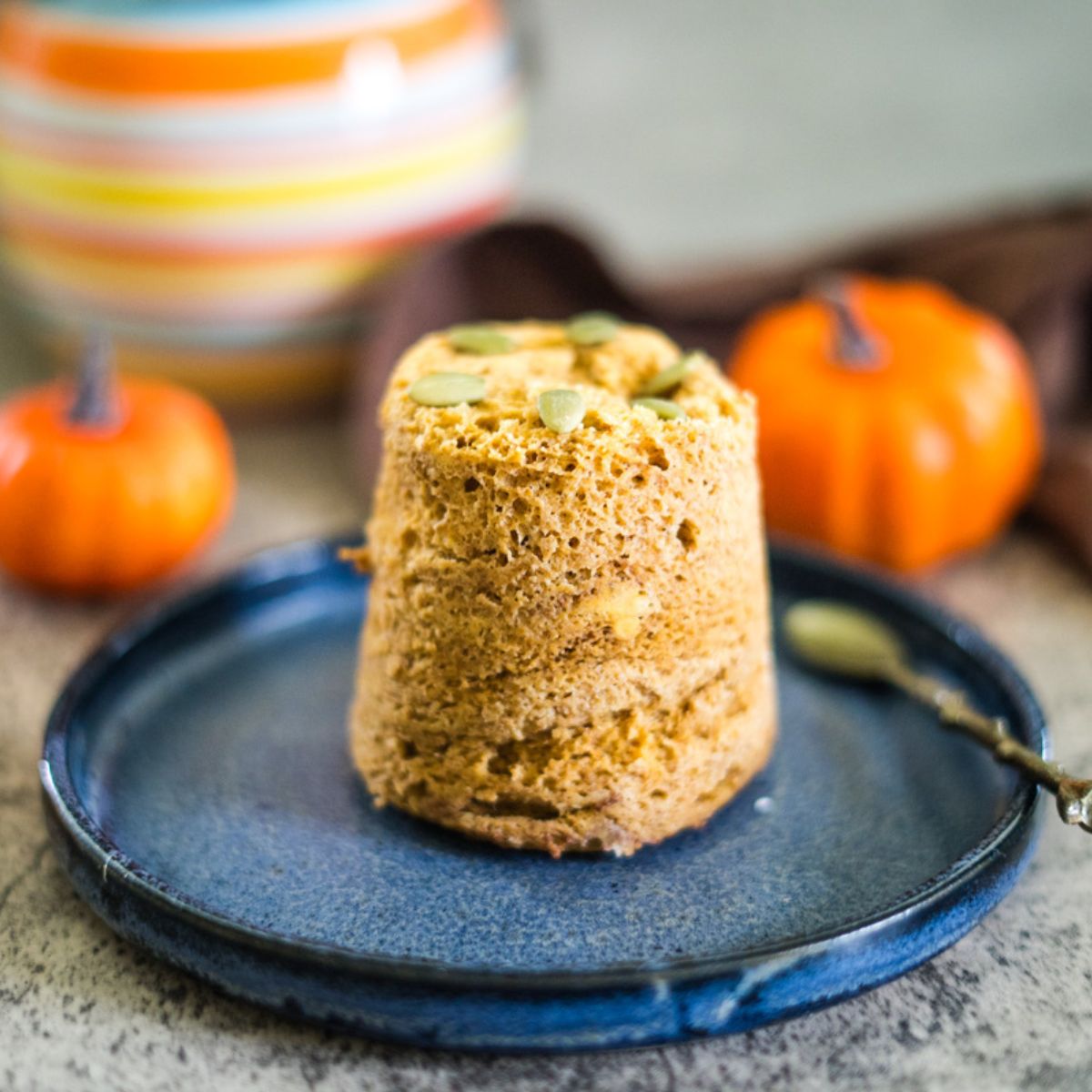 A mug cake on a blue plate, topped with seeds, with small decorative pumpkins and a striped jar in the background.