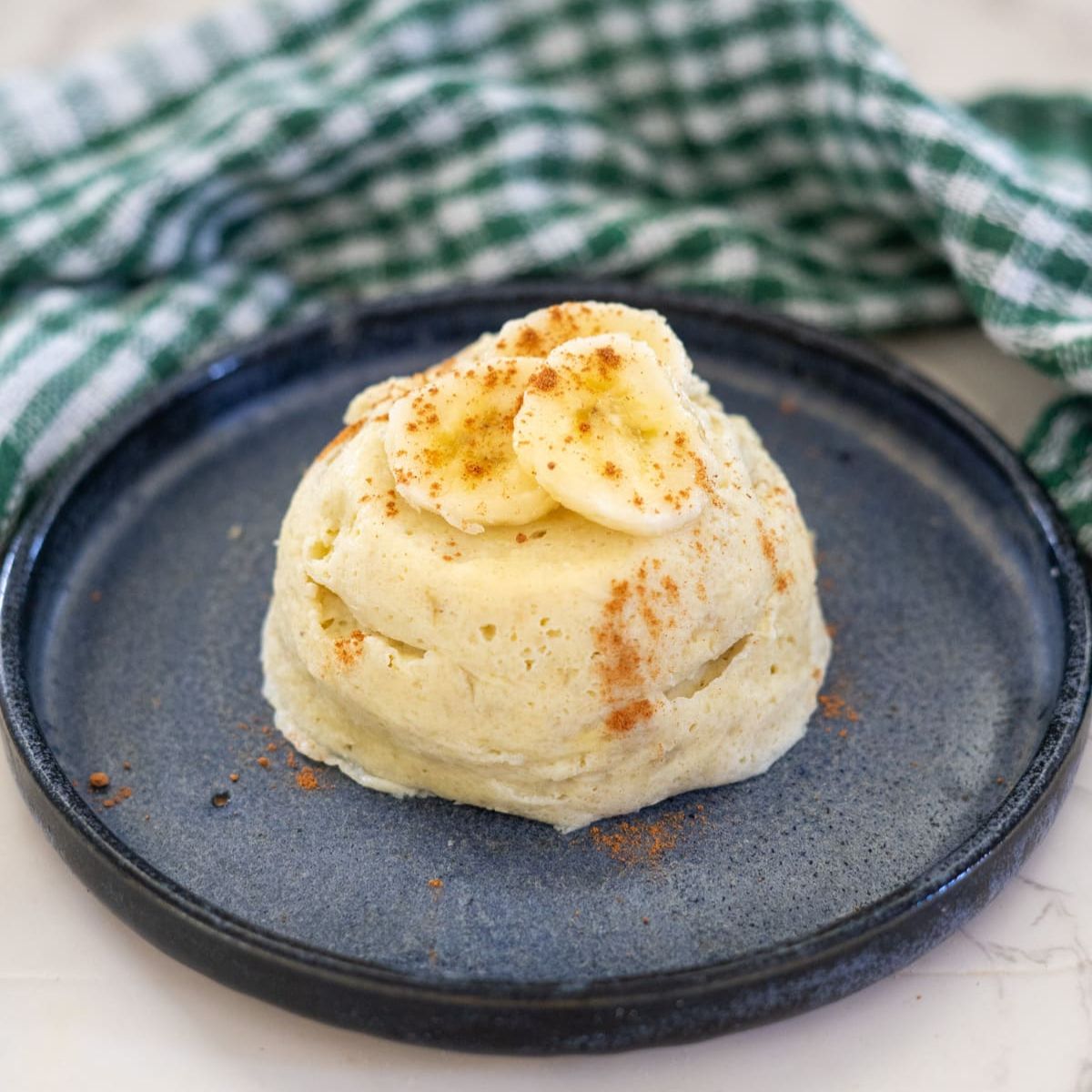A banana cake topped with sliced bananas and sprinkled cinnamon on a blue plate, with a green and white checkered cloth in the background.