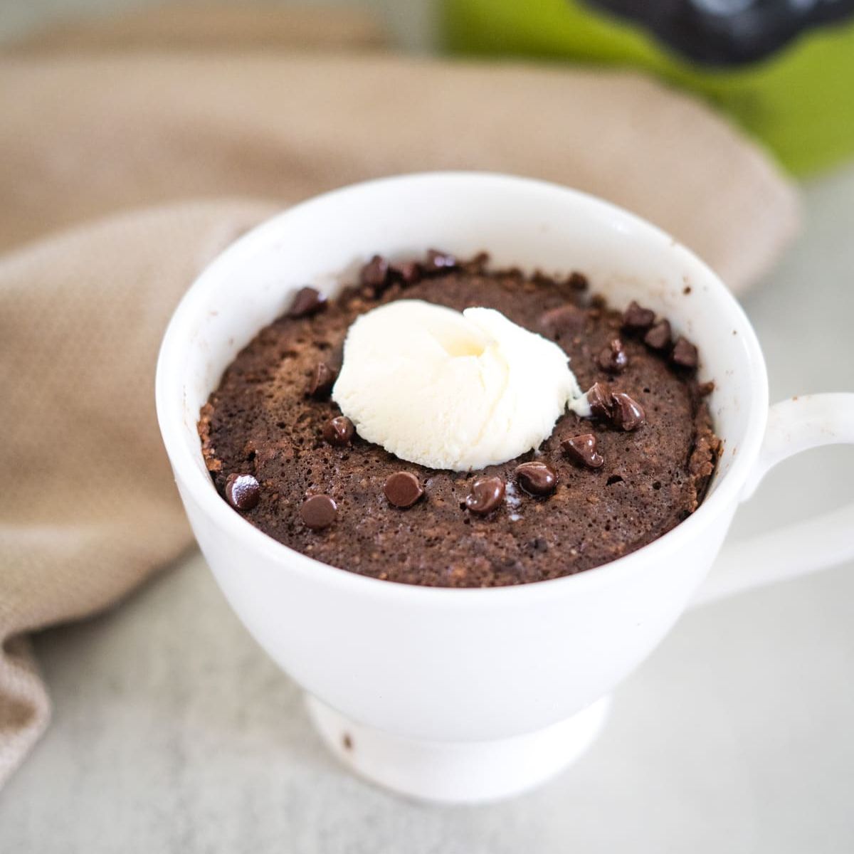 Chocolate mug cake with chocolate chips and a dollop of whipped cream in a white cup on a gray surface.