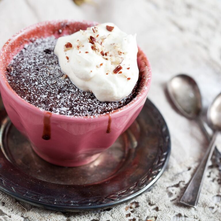 Chocolate mug cake with powdered sugar and whipped cream in a pink cup, placed on a metal saucer. Two spoons lie beside it on a lace tablecloth.