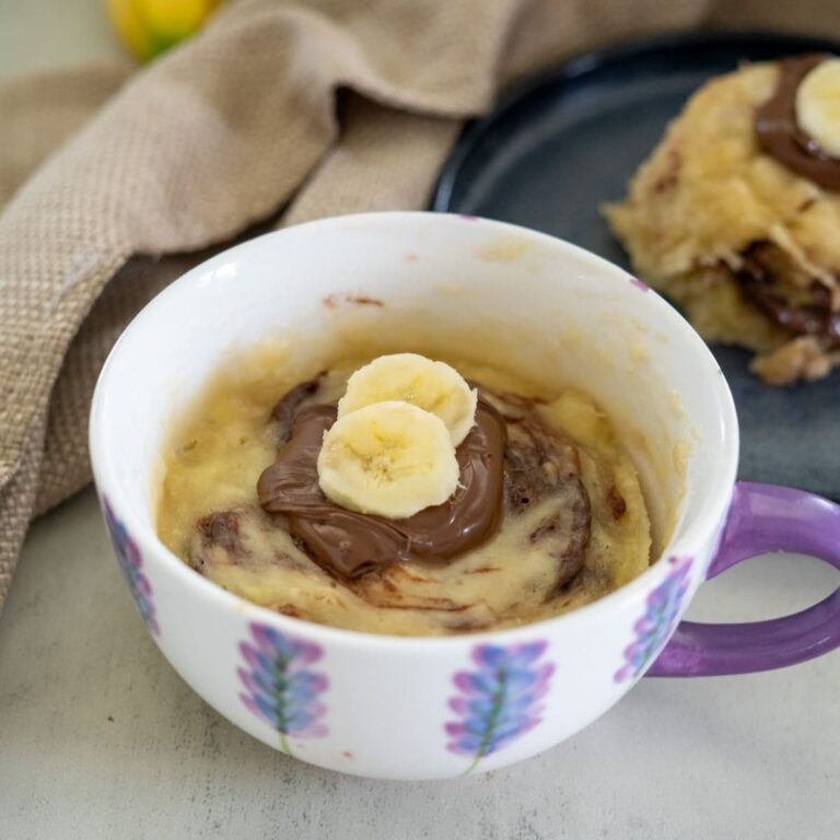 A mug cake topped with banana slices and chocolate spread, served in a white cup with floral patterns. Another piece is on a dark plate nearby.