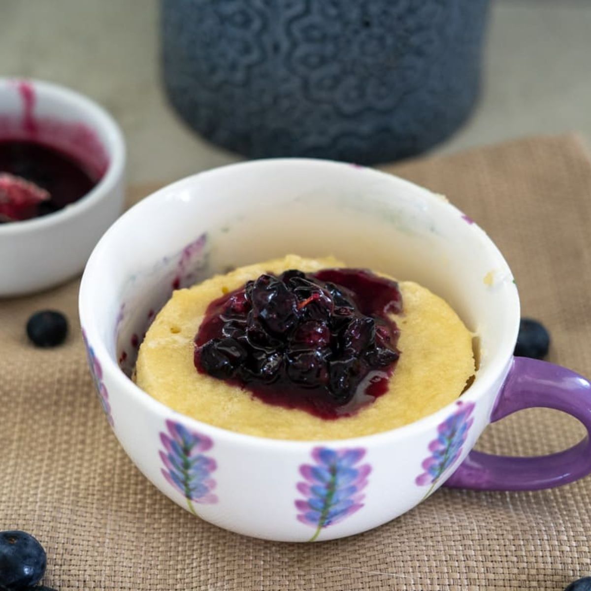 A mug cake topped with blueberry sauce in a floral cup with a purple handle, placed on a woven mat.