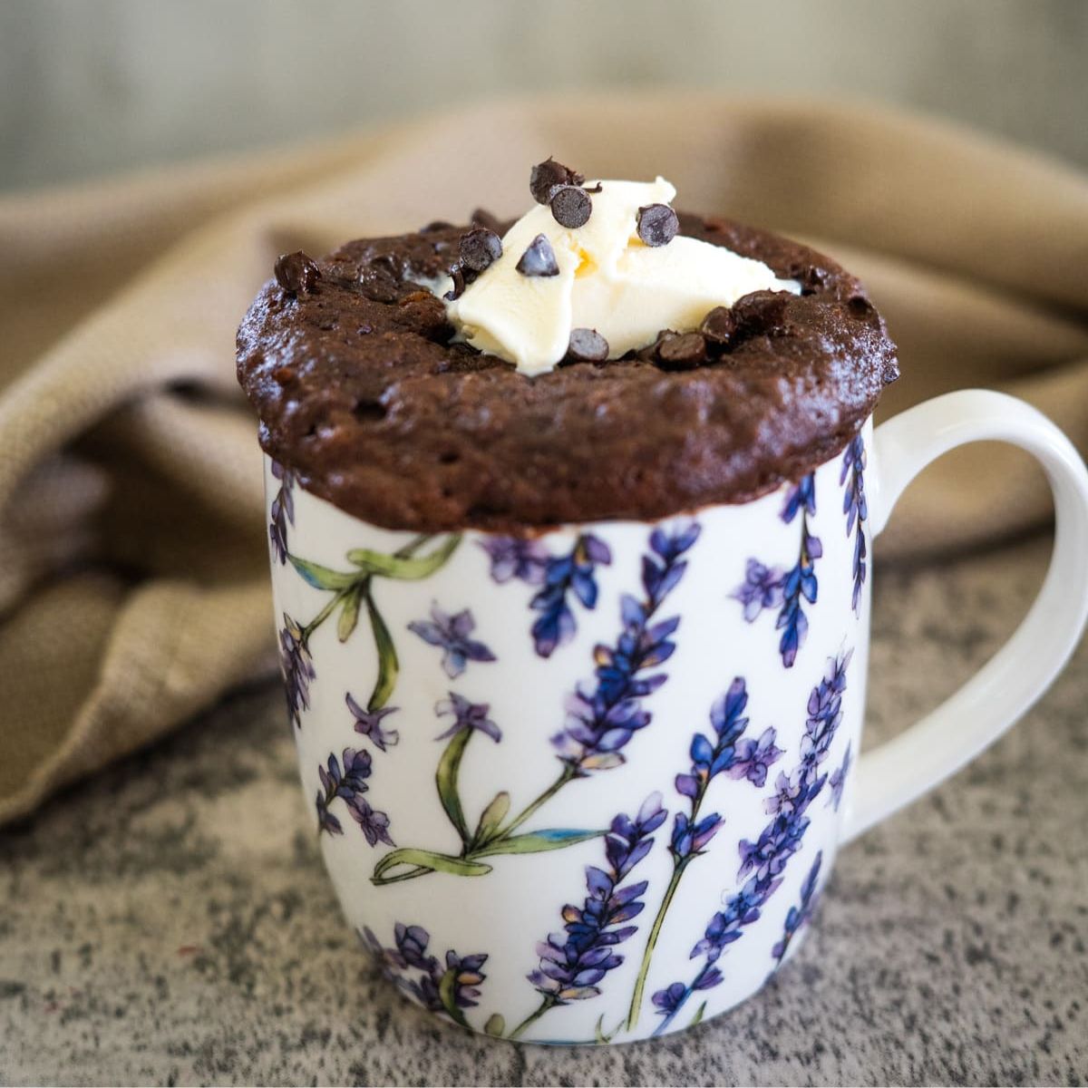 Chocolate mug cake in a floral mug, topped with whipped cream and chocolate chips, on a textured surface with a beige cloth in the background.