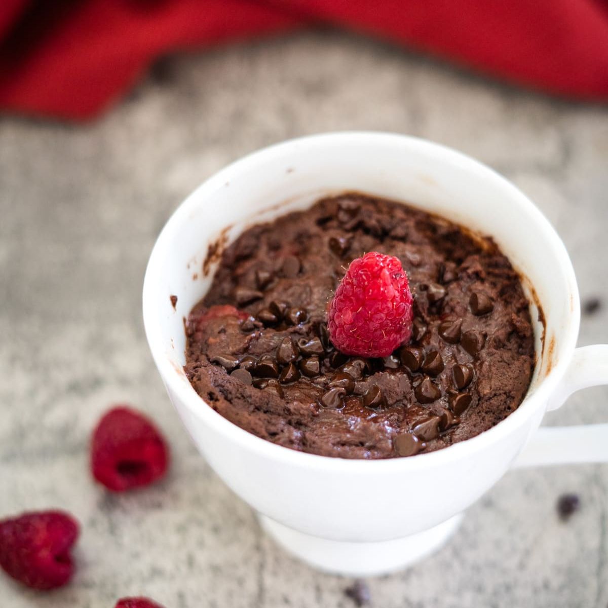 Chocolate mug cake with chocolate chips topped with a raspberry, in a white cup, placed on a gray surface with additional raspberries nearby.