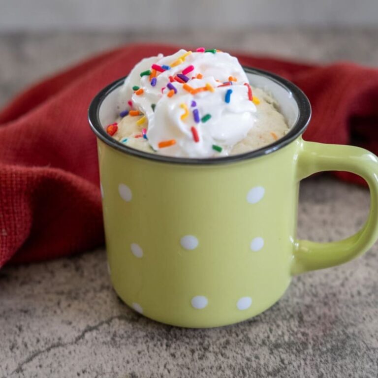 A yellow polka dot mug filled with a dessert, topped with whipped cream and colorful sprinkles, placed on a textured surface with a red cloth in the background.