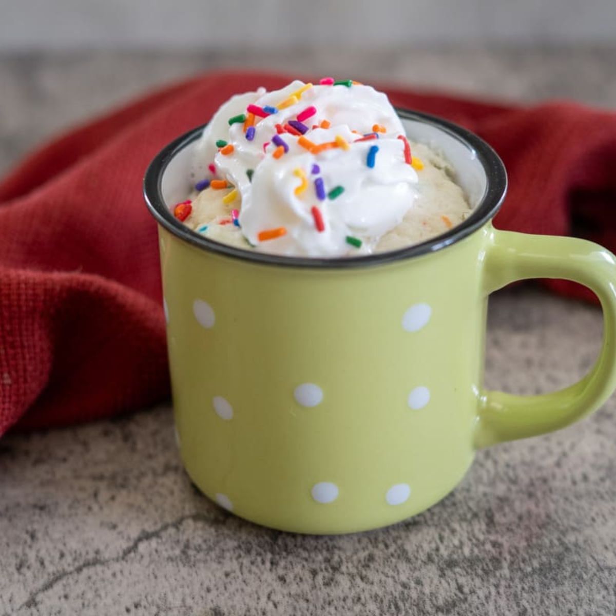 A yellow polka dot mug filled with a dessert, topped with whipped cream and colorful sprinkles, placed on a textured surface with a red cloth in the background.
