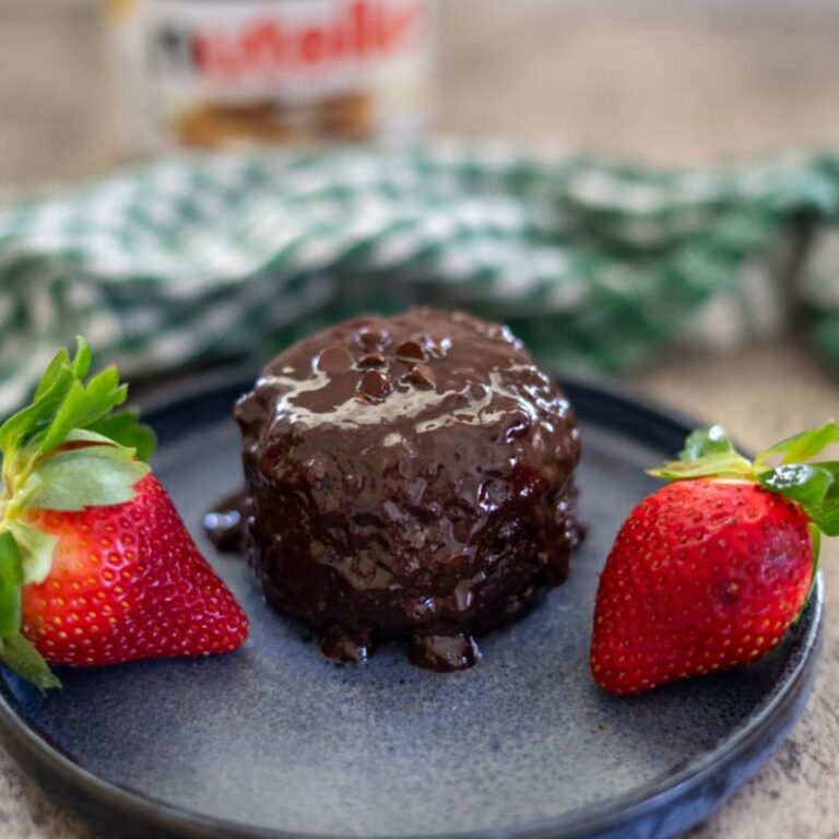 Chocolate lava cake on a black plate with two strawberries, set on a wooden surface. Nutella jar in the blurry background.