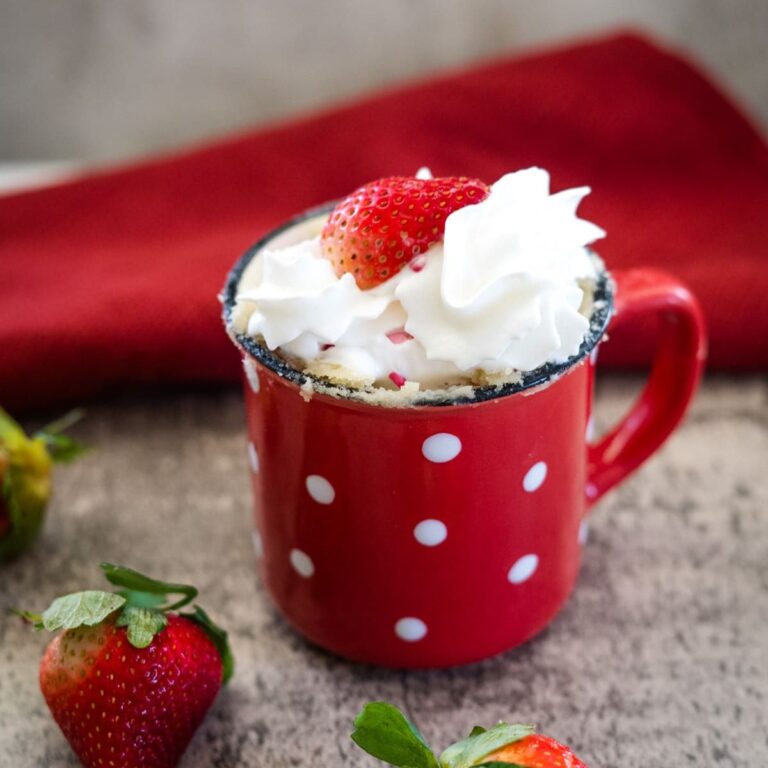 A red mug with white polka dots, filled with whipped cream and topped with a strawberry, is placed on a gray surface with more strawberries nearby. A red cloth is in the background.