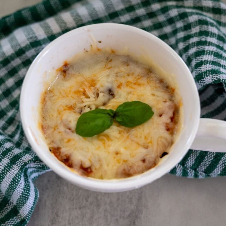 A mug of lasagna topped with melted cheese and a basil leaf, placed on a gray surface with a green and white checkered cloth nearby.