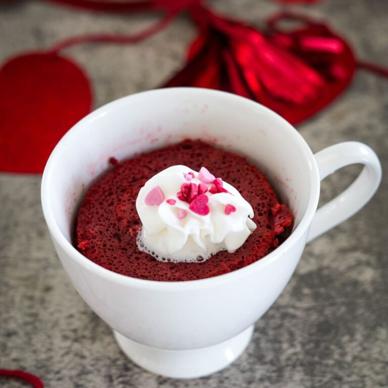 A white mug with a red velvet microwave cake topped with whipped cream and pink heart sprinkles, placed on a gray surface. Red decorations are slightly visible in the background.