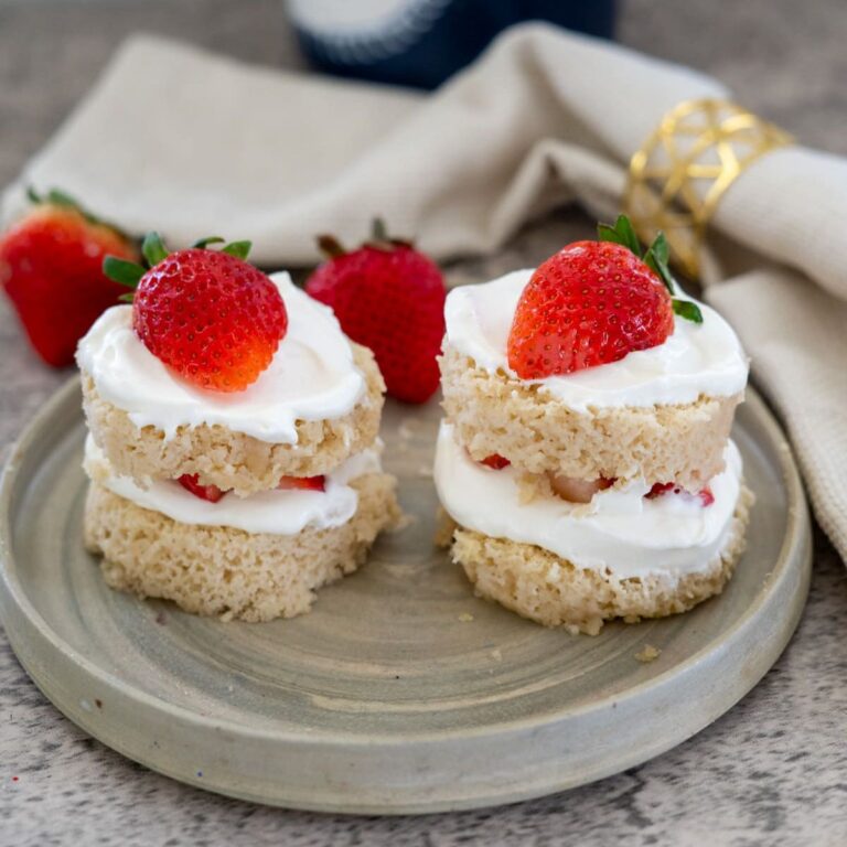 Two mini layered cakes with whipped cream and strawberries on top, placed on a round ceramic plate. A cloth napkin and whole strawberries are in the background.