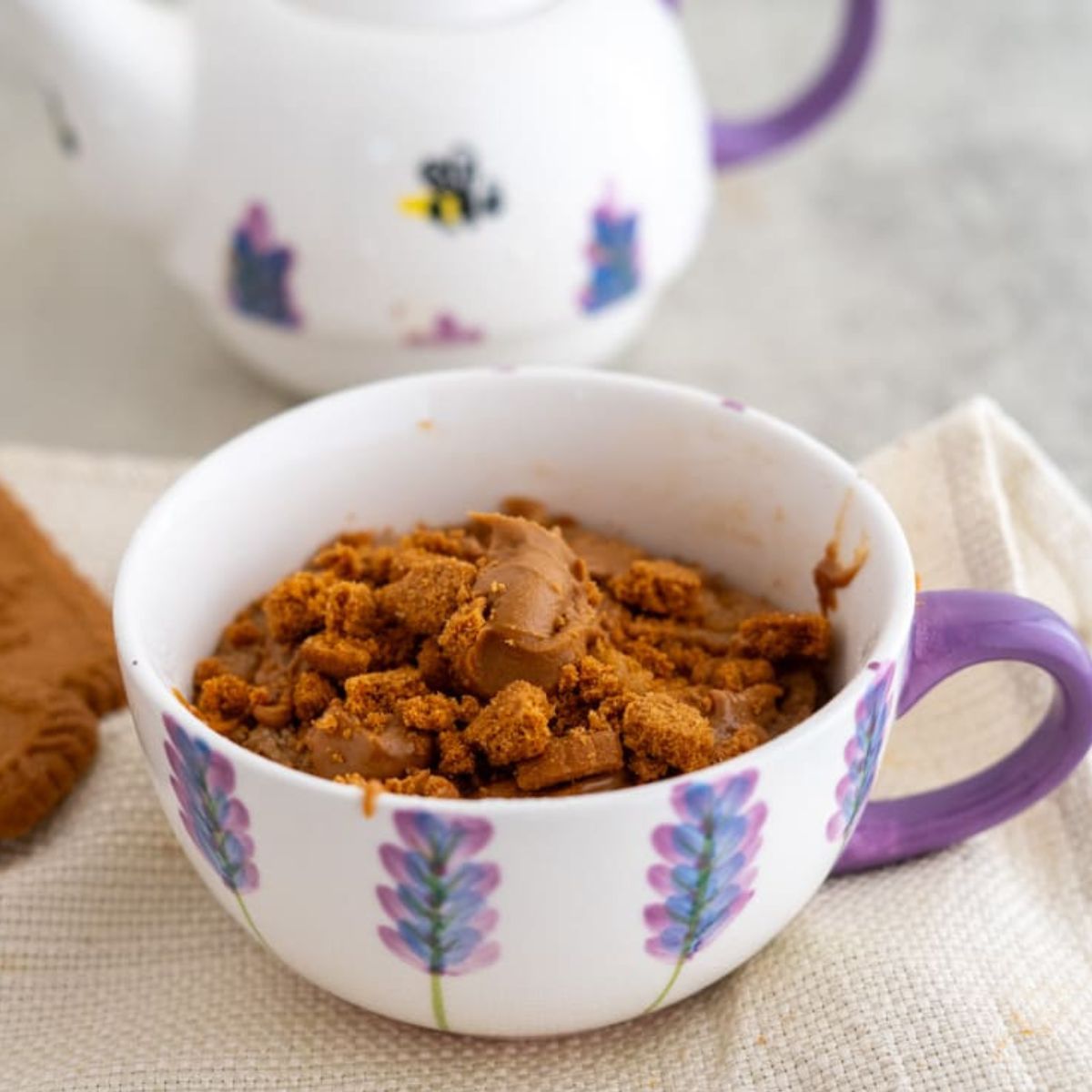 A cup with purple floral design contains brown crumbly dessert with a piece of chocolate on top; a teapot is in the background.