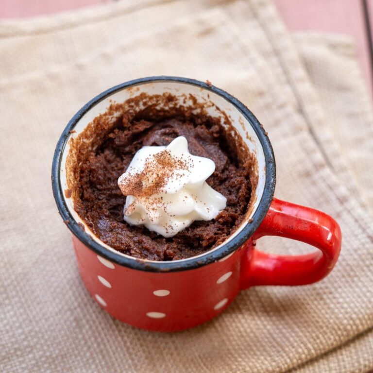 A red polka dot mug filled with a delicious Nutella mug cake recipe, topped with whipped cream and a sprinkle of cocoa, sits on a beige cloth.