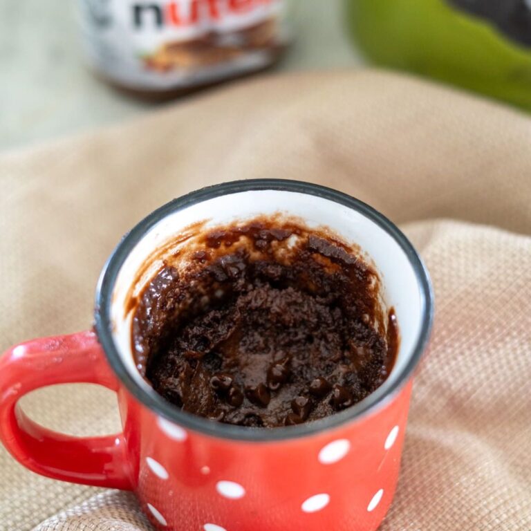 Red mug with white polka dots containing a partially eaten chocolate mug cake. Nutella jar blurred in the background.