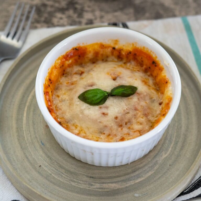 A baked dish in a white ramekin topped with melted cheese and a basil leaf, placed on a round plate next to a fork.