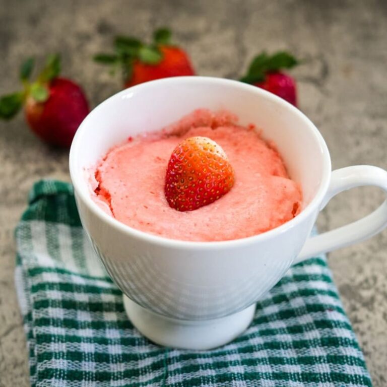 A white mug with strawberry cake topped by a single strawberry, placed on a green checkered cloth. Three strawberries are in the background.