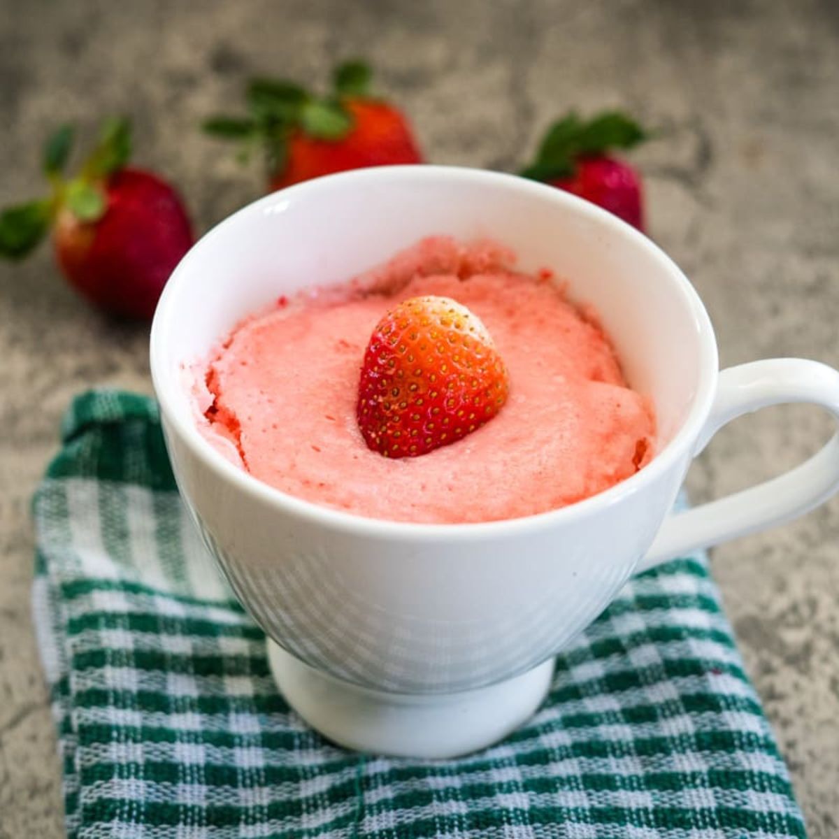 A white mug with strawberry cake topped by a single strawberry, placed on a green checkered cloth. Three strawberries are in the background.