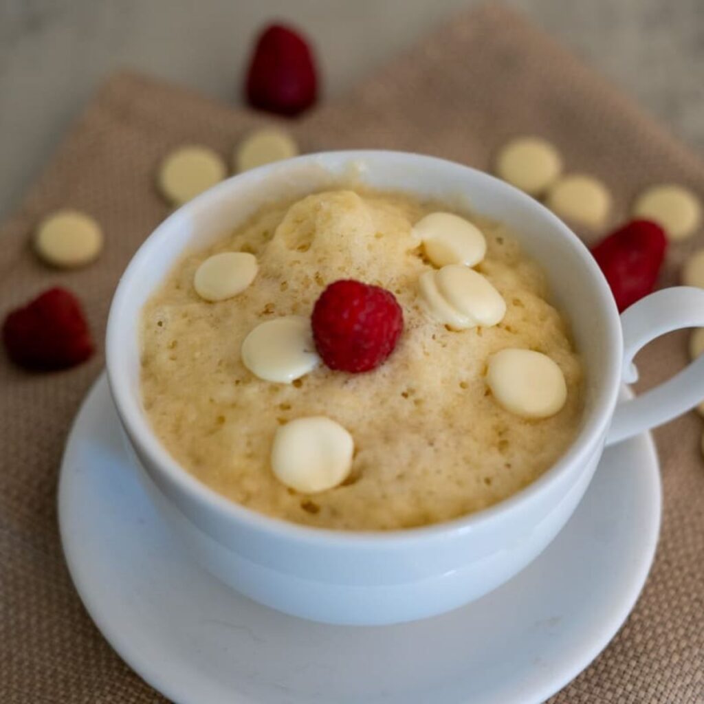 A white mug on a saucer contains a vanilla mug cake topped with white chocolate chips and a single raspberry. Additional raspberries and chocolate chips are scattered on a brown cloth.