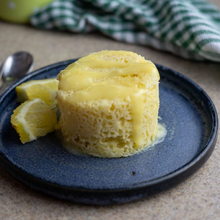 A small, round lemon mug cake topped with lemon glaze sits on a blue plate, with lemon wedges on the side and a green checkered cloth in the background.