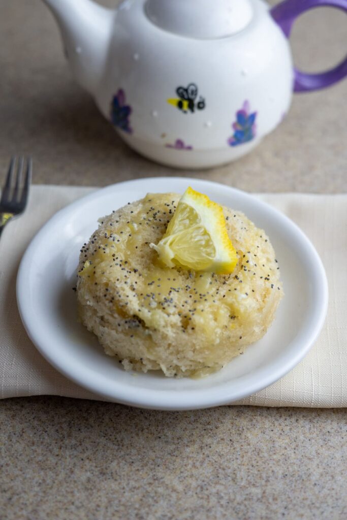 A small round lemon poppy seed cake topped with a lemon wedge on a white plate, with a teapot in the background.
