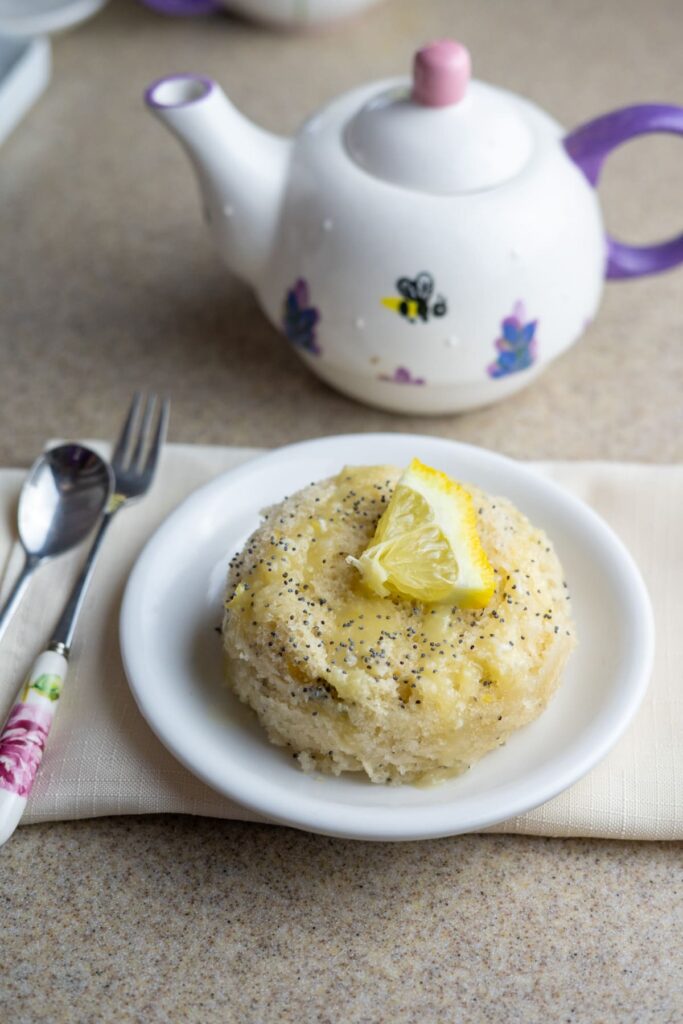A small lemon poppy seed mug cake topped with a lemon wedge sits on a white plate, with a spoon, fork, and decorated teapot in the background.
