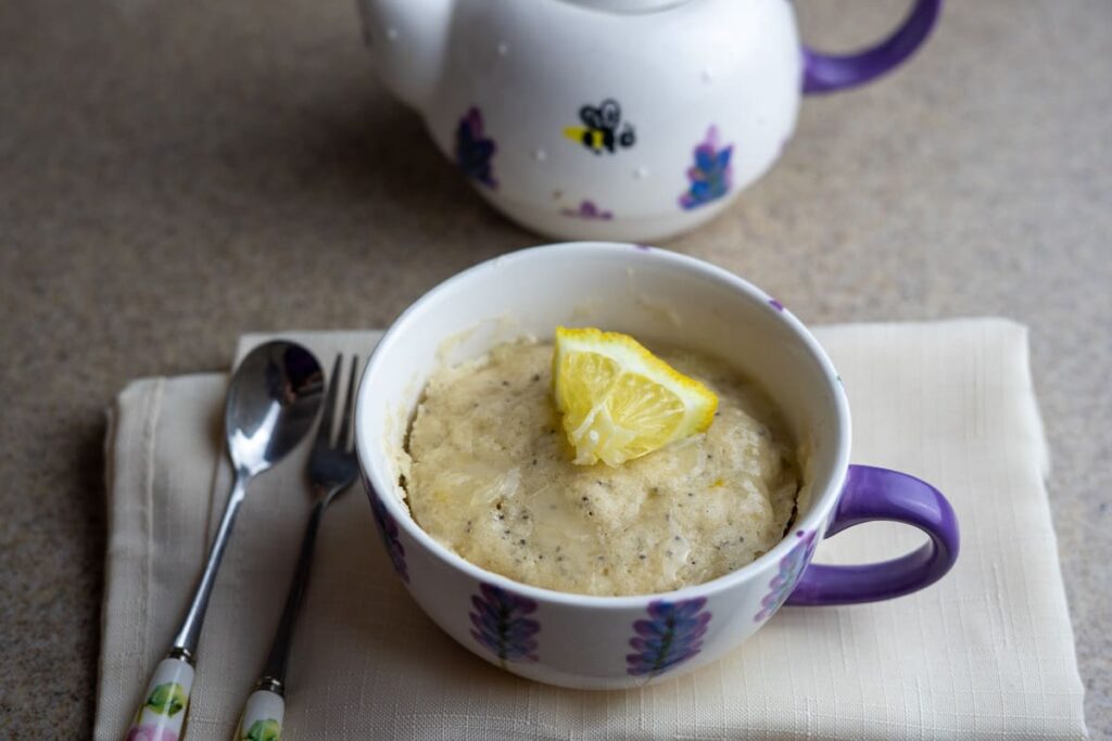 A ceramic mug with a baked mug cake, topped with a lemon wedge, sits on a napkin next to two spoons, with a teapot in the background.