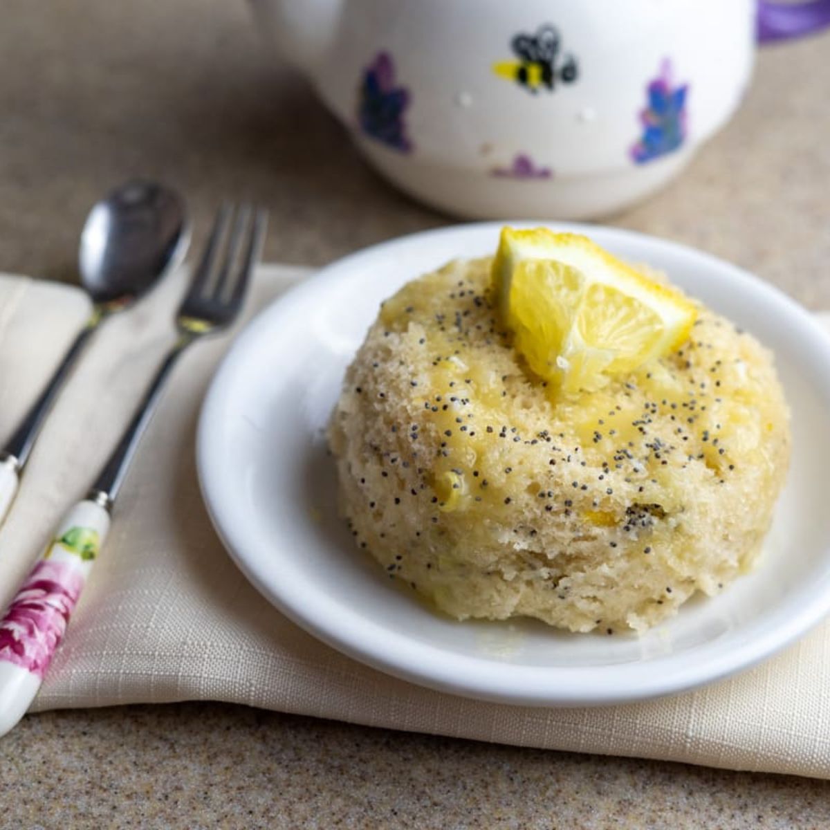 A lemon poppy seed mug cake topped with a lemon wedge is served on a white plate, next to a fork and spoon on a napkin, with a teapot in the background.
