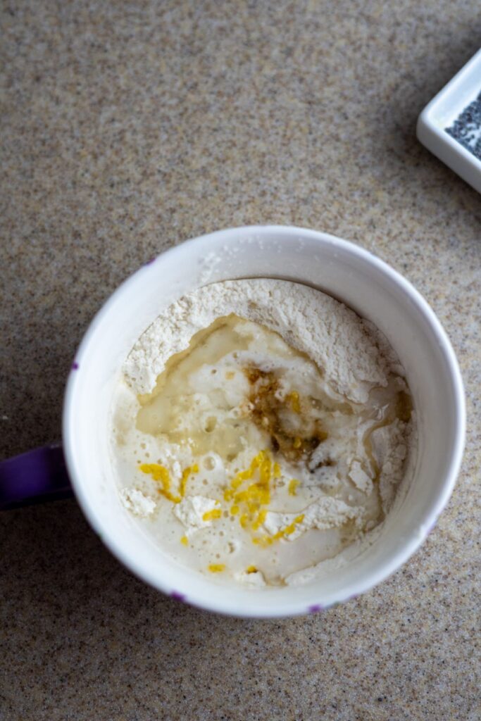 A mug filled with flour, zest, liquid ingredients, and vanilla extract on a countertop, ready to be mixed.
