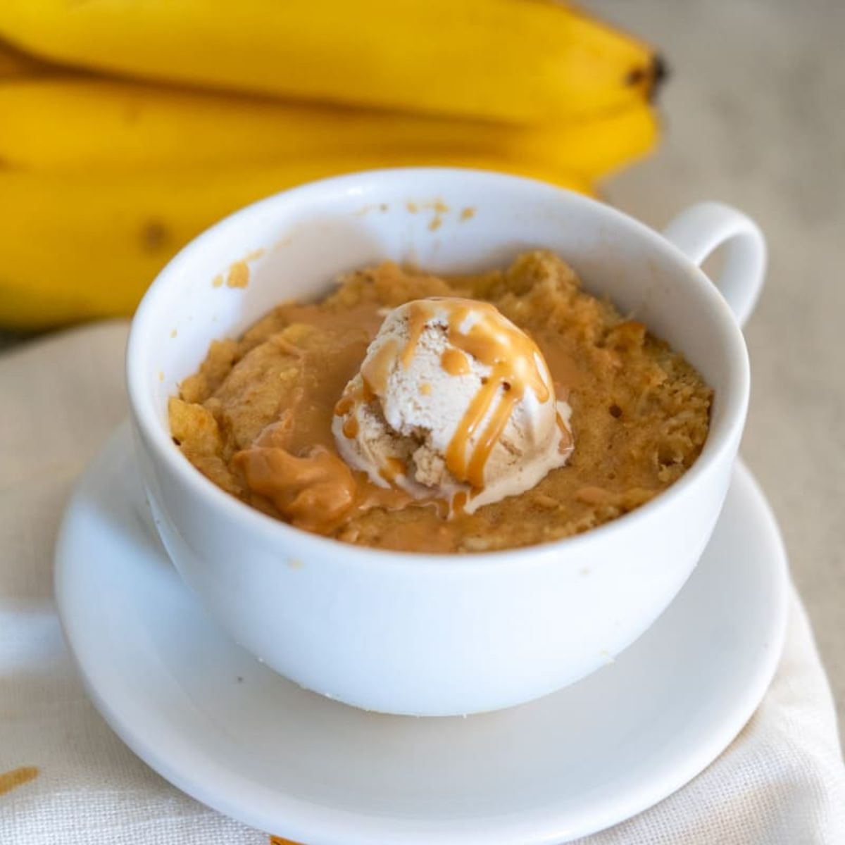 A white cup filled with banana mug cake, topped with a scoop of ice cream and caramel drizzle, sits on a white saucer with bananas in the background.