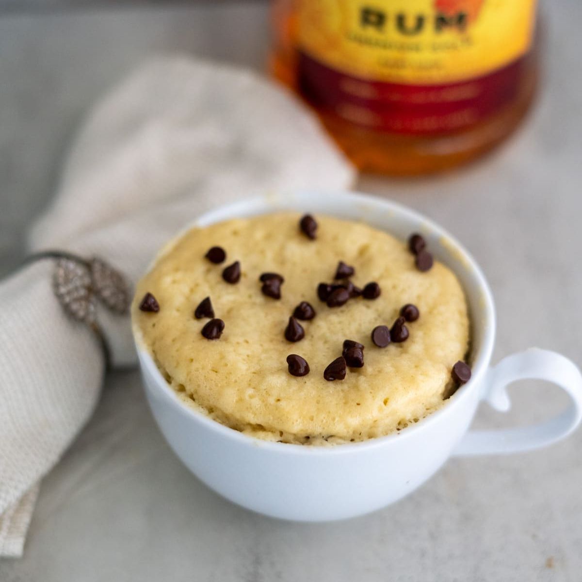 A white cup filled with a microwave mug cake topped with chocolate chips sits on a countertop next to a beige napkin and a bottle of rum in the background.