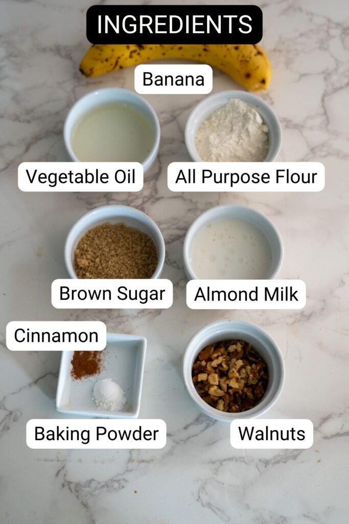 Various baking ingredients are arranged in small bowls on a marble surface, labeled as banana, vegetable oil, all purpose flour, brown sugar, almond milk, cinnamon, baking powder, and walnuts.