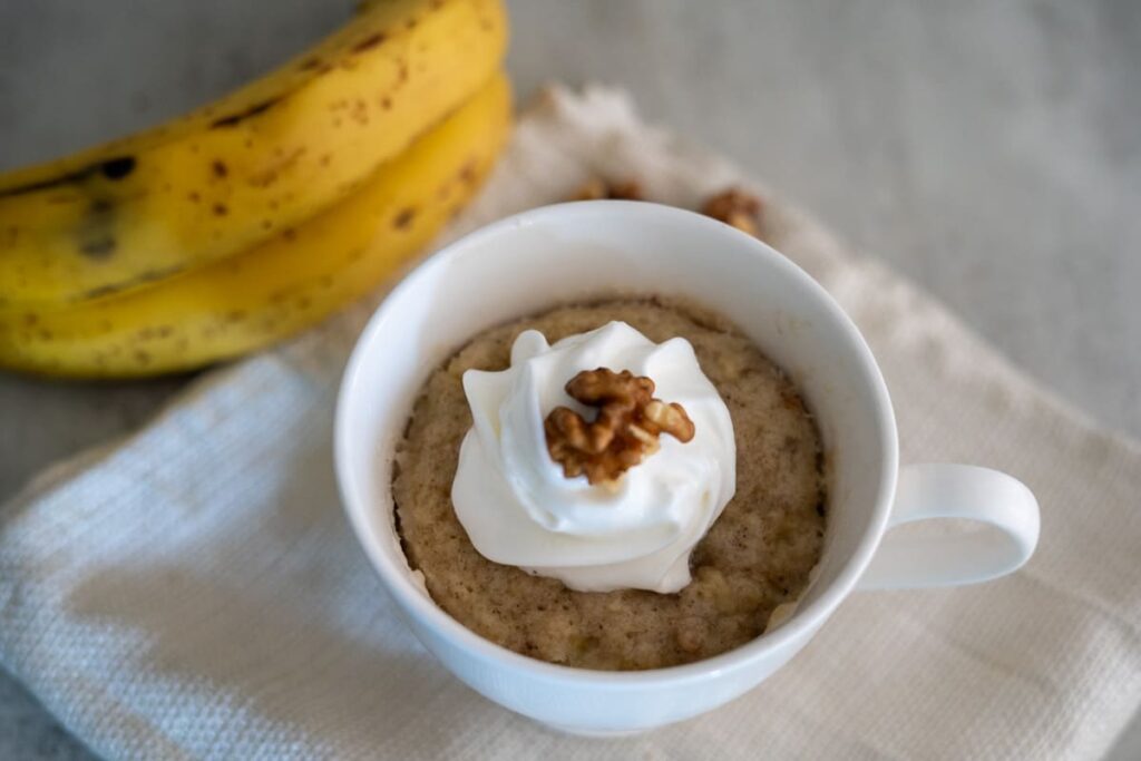 A white cup filled with oatmeal topped with whipped cream and a walnut, with bananas in the background on a light surface.