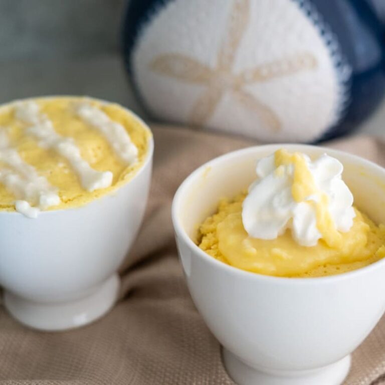 Two white cups filled with yellow mug cake, each topped with whipped cream, are placed on a beige cloth with a blue and white decorative object in the background.