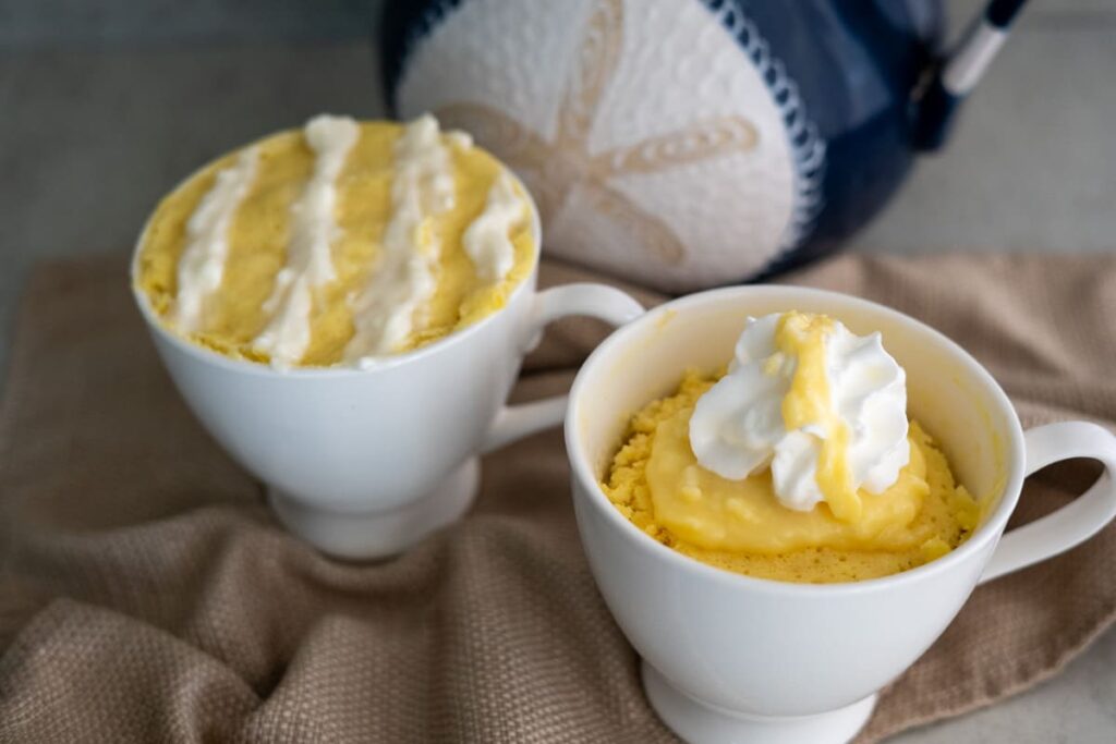 Two white mugs filled with individual yellow mug cakes, each topped with whipped cream, are placed on a beige cloth with a blue and white ceramic jar in the background.
