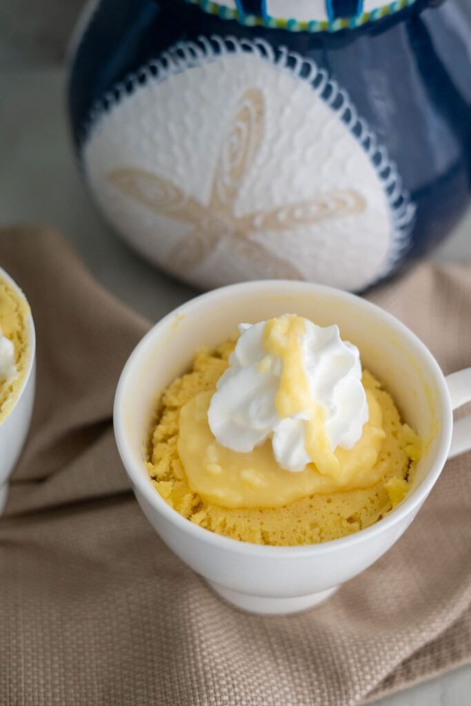 A white mug filled with a yellow cake, topped with a dollop of custard and whipped cream, sits on a beige cloth with a blue and white decorative object in the background.