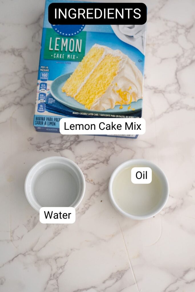 A box of lemon cake mix, a small bowl of water, and a small bowl of oil are arranged on a marble surface.