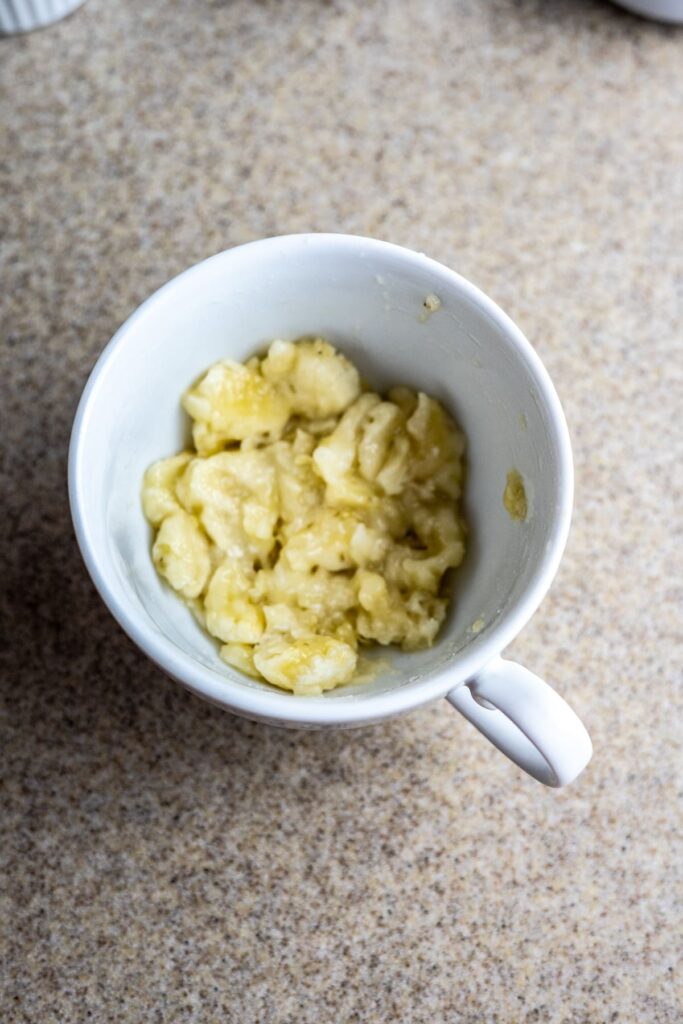 A white mug containing mashed bananas sits on a speckled countertop.