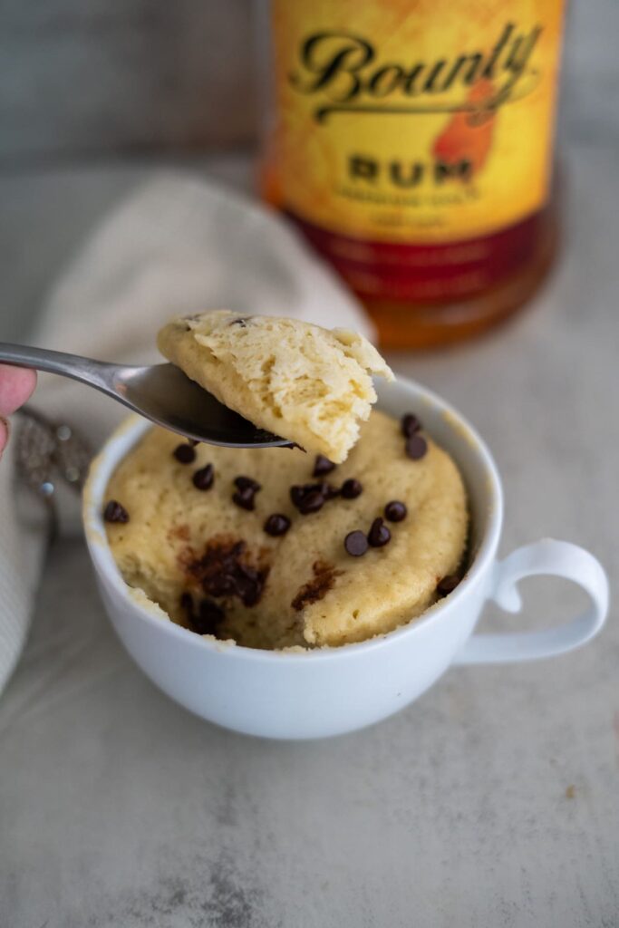 A white cup filled with a banana rum mug cake topped with chocolate chips, with a spoon holding a bite above it. A bottle of Bounty rum is blurred in the background.