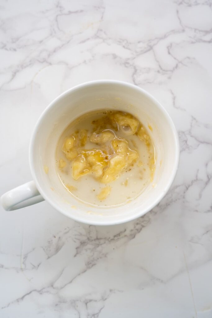 A white mug on a marble surface containing mashed banana mixed with liquid.