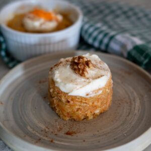 A small round carrot cake topped with white frosting and a walnut sits on a beige plate, with another dessert and a green checkered cloth in the background.