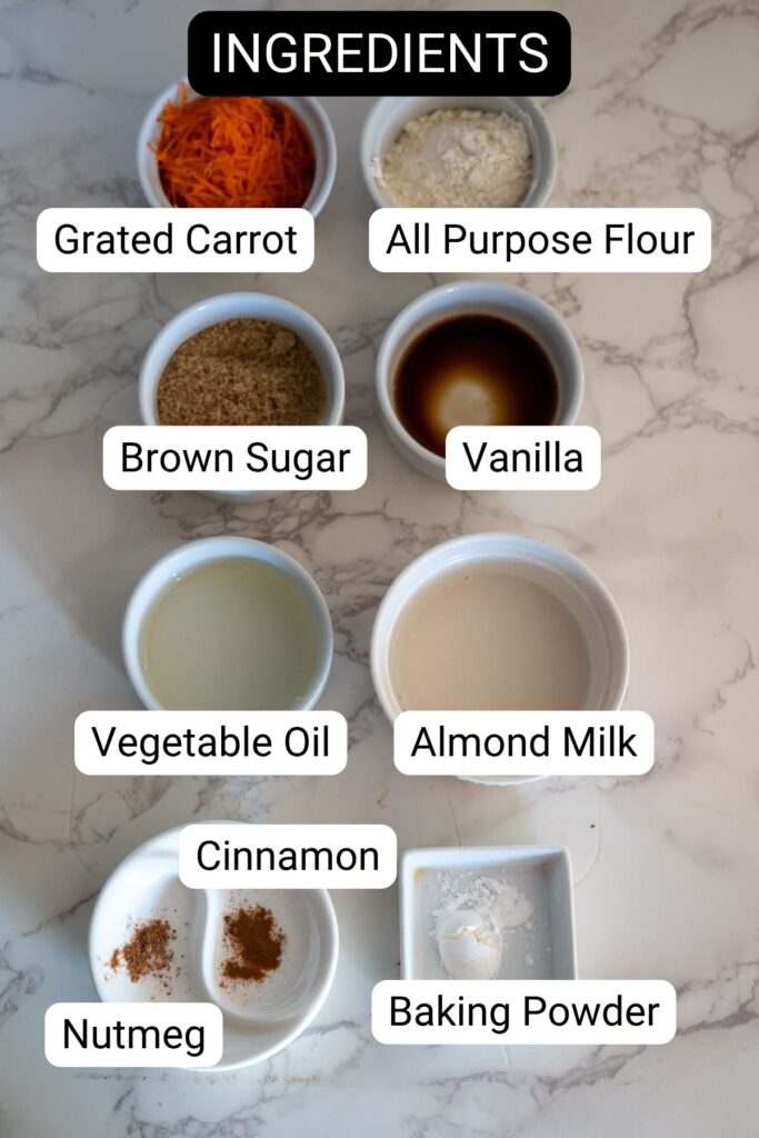Top-down view of labeled bowls containing grated carrot, flour, brown sugar, vanilla, vegetable oil, almond milk, nutmeg, cinnamon, and baking powder on a marble surface.