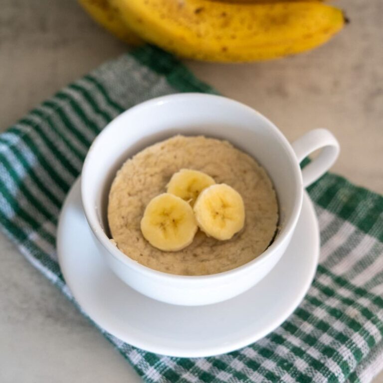A white cup with banana mug cake topped with banana slices, placed on a matching saucer and a green striped cloth, with a banana in the background.
