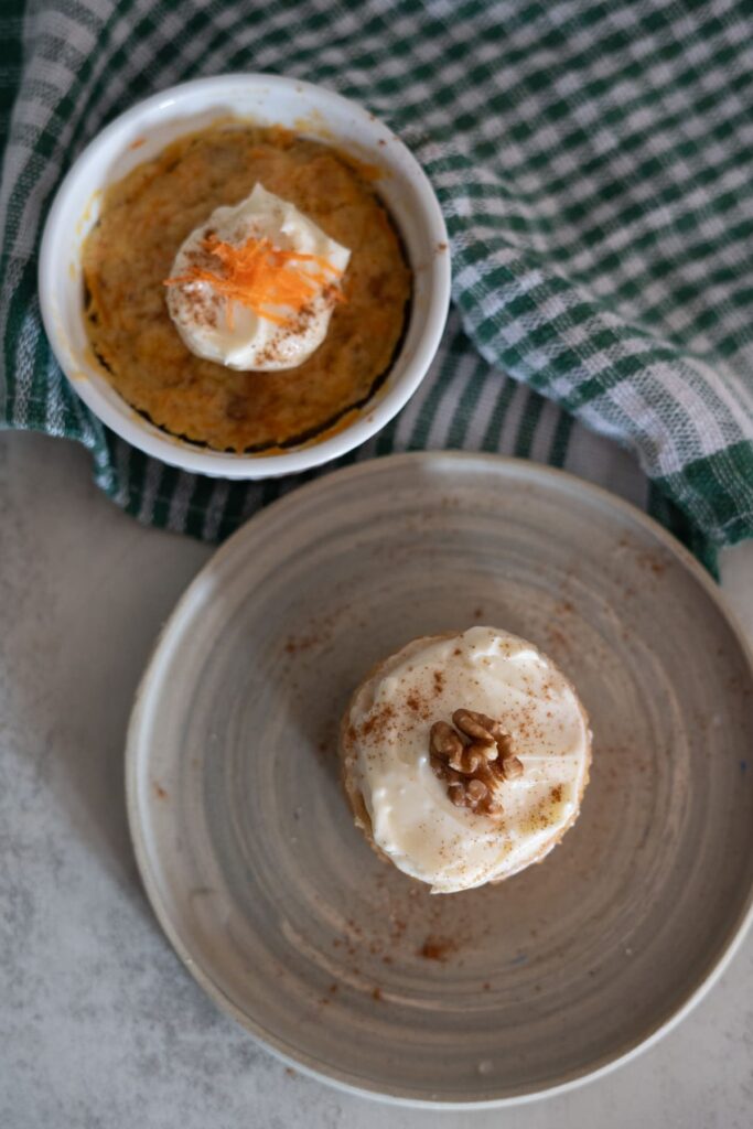 A small baked dessert in a ramekin and a frosted cupcake on a plate, both topped with cream and garnishes, next to a green checkered cloth.