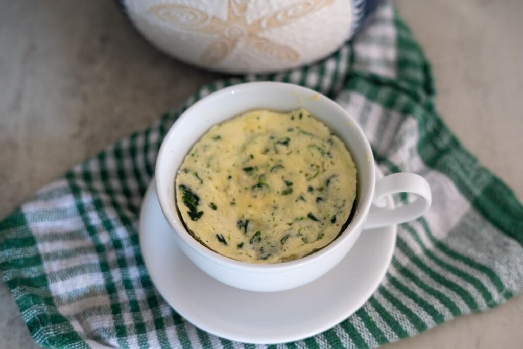 A cup filled with a cooked egg mixture containing herbs, placed on a white saucer atop a green and white striped cloth.