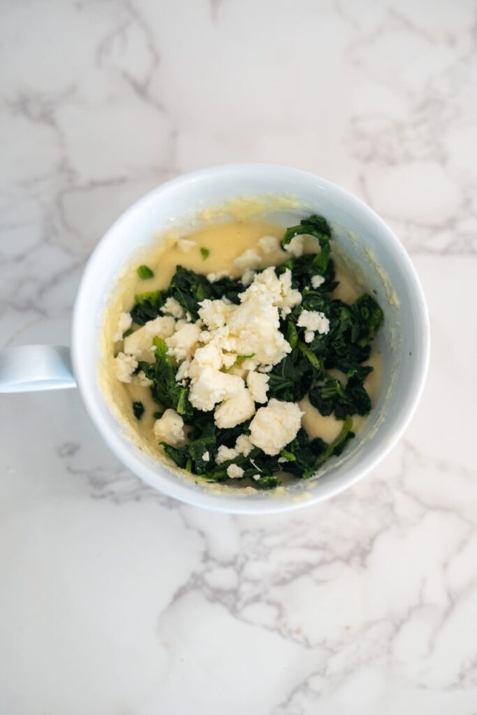 A white mug containing a mixture of batter, chopped spinach, and crumbled cheese sits on a white marble surface.