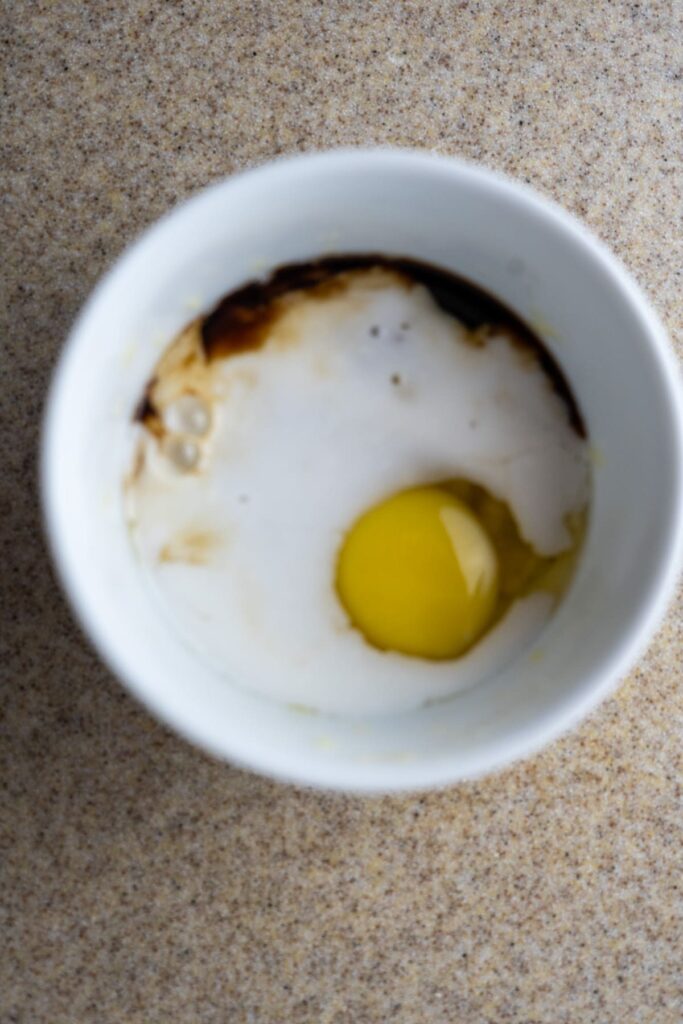 A cracked raw egg, milk, and a dark liquid, possibly vanilla extract, in a white bowl on a speckled countertop.