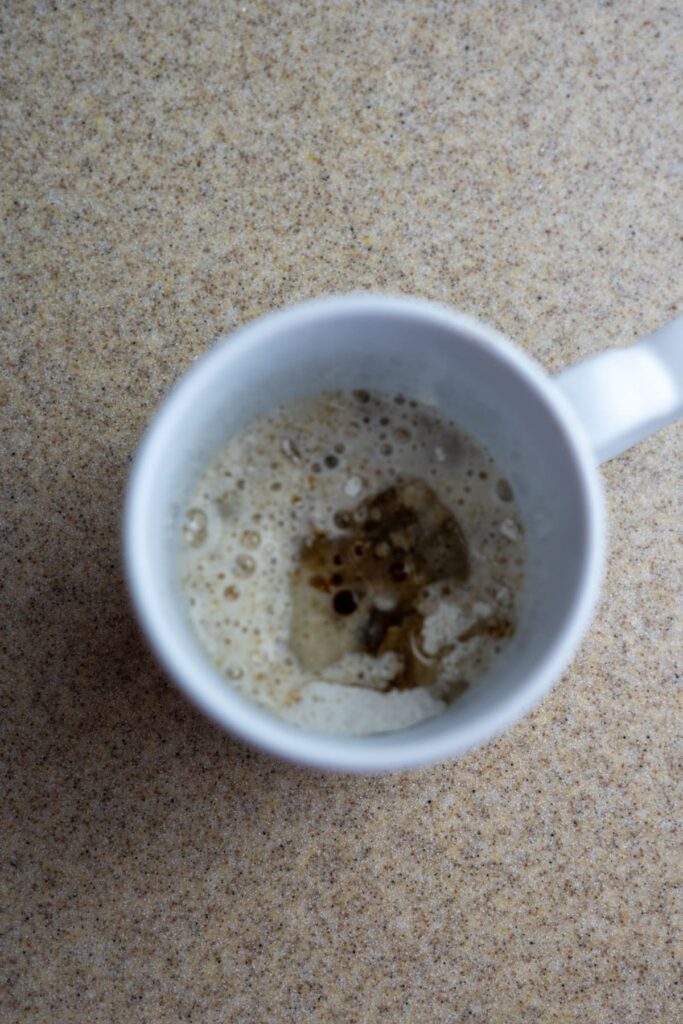 White mug with light brown frothy coffee residue inside, seen from above, placed on a speckled beige countertop.