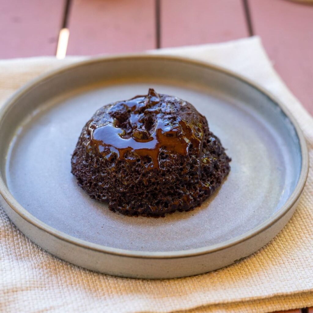 A chocolate lava cake sits on a ceramic plate, topped with melted chocolate sauce, placed on a light-colored cloth.
