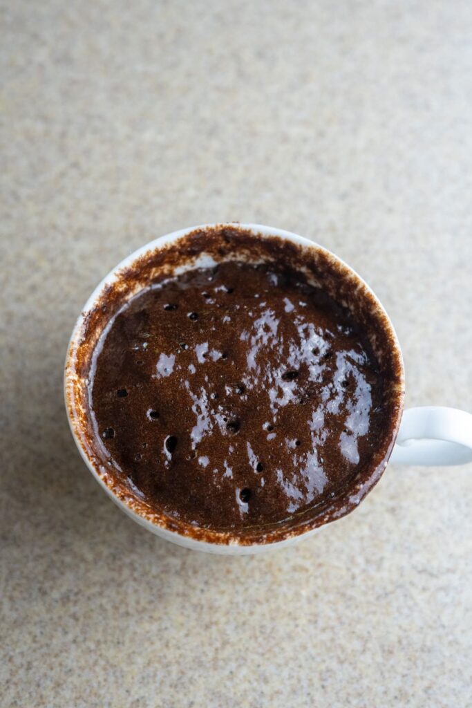 A white mug containing a partially cooked keto brownie mug cake mixture with small holes on the surface, placed on a beige countertop.