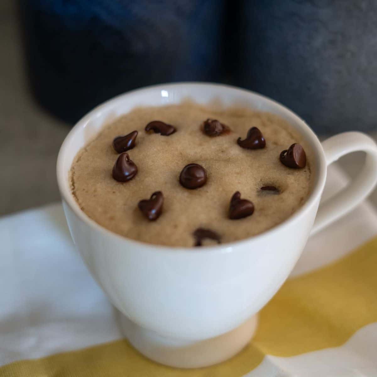 A white mug filled with a chocolate chip mug cake, topped with several chocolate chips, sits on a striped cloth.